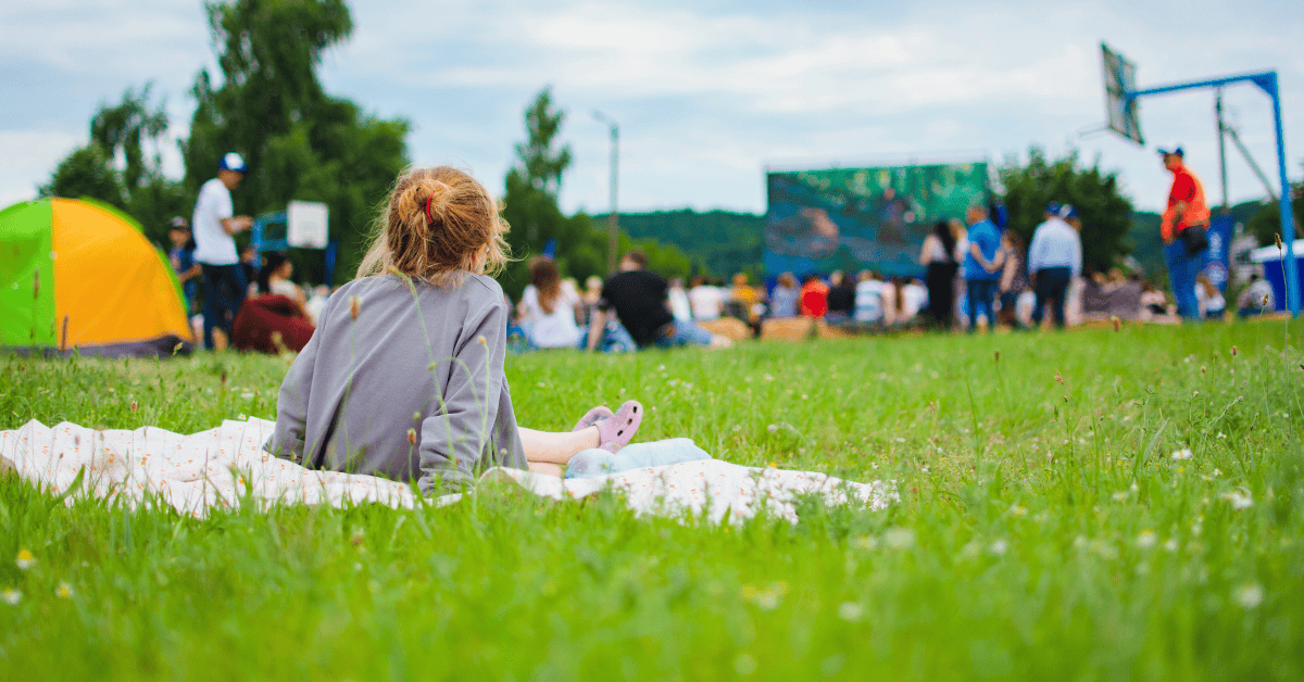 A child sitting alone on a blanket in the grass watching groups of people in the distance.